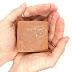 Red clay soap bar in hands, tan-brown with suds