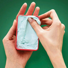 Hands holding open hair styling matte cream in small red container with pale blue gel
